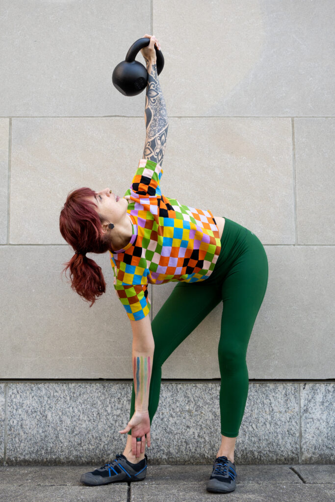 Sarah, in green workout leggings and a rainbow checkered shirt, does a kettlebell windmill exercise by a stone wall.