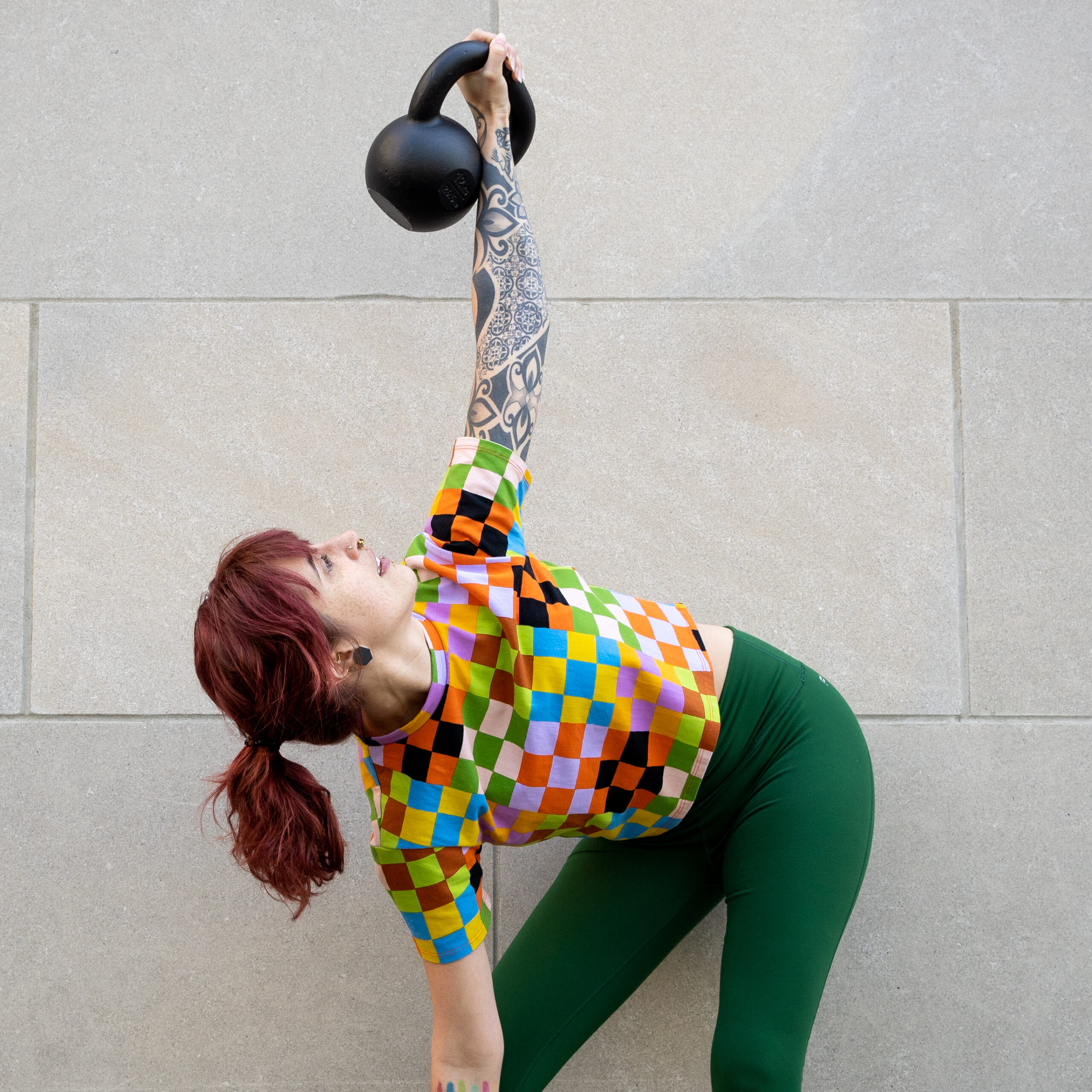Sarah, in green workout leggings and a rainbow checkered shirt, does a kettlebell windmill exercise by a stone wall. Should I lift weights?