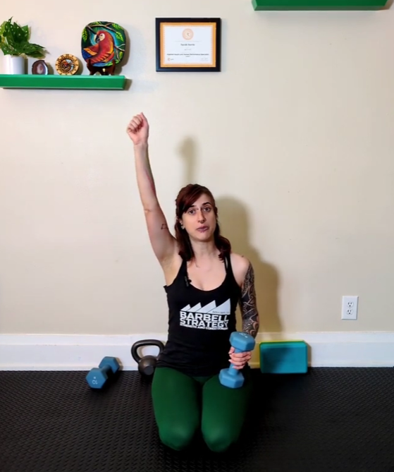 Sarah demonstrates an overhead press while holding a blue dumbbell